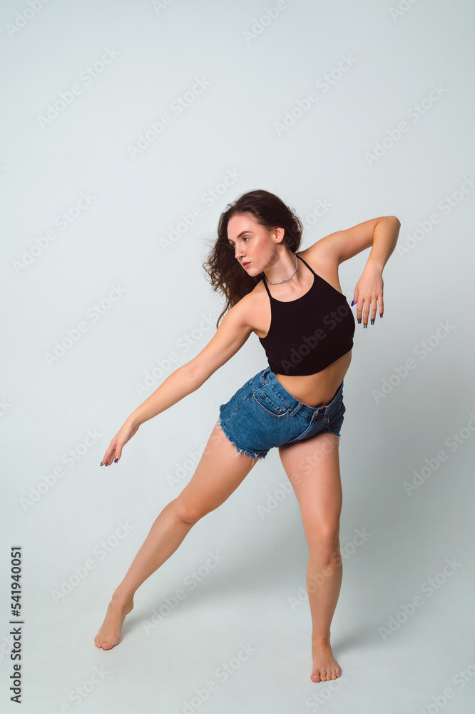 Obraz premium young beautiful woman with brown hair in a black top and short denim shorts dances barefoot on a white background. Full length portrait