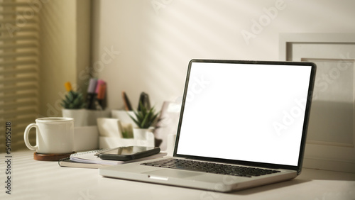 Laptop computer with empty display, coffee cup, picture frame and stationery on white table.