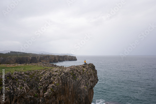 Large cliffs on the Asturian coast with an adult woman in the background looking towards the Cantabrian Sea on a rainy day in Asturias, Spain.