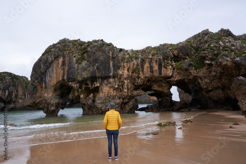 Adult person with yellow jacket on the beach in front of the beautiful rock formations with caves of a tourist protected area on the eastern coast of Asturias, Spain. Famous beach of Cuevas del Mar.