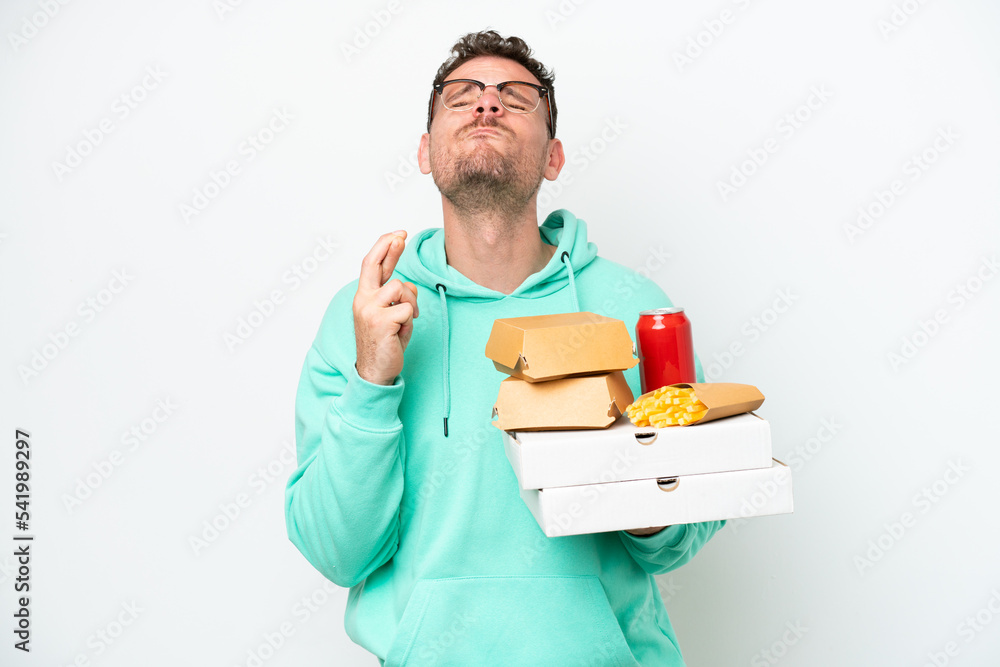 Young caucasian man holding fast food isolated on white background with fingers crossing and wishing the best Young caucasian man holding fast food isolated on white background with fingers crossing and wishing the best