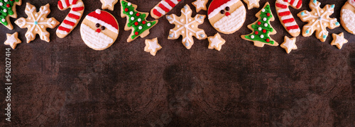 Cute Christmas cookie top border. Overhead view on a dark stone banner background with copy space. Holiday baking concept. Santa Claus, tree, snowflakes, candy canes, stars.
