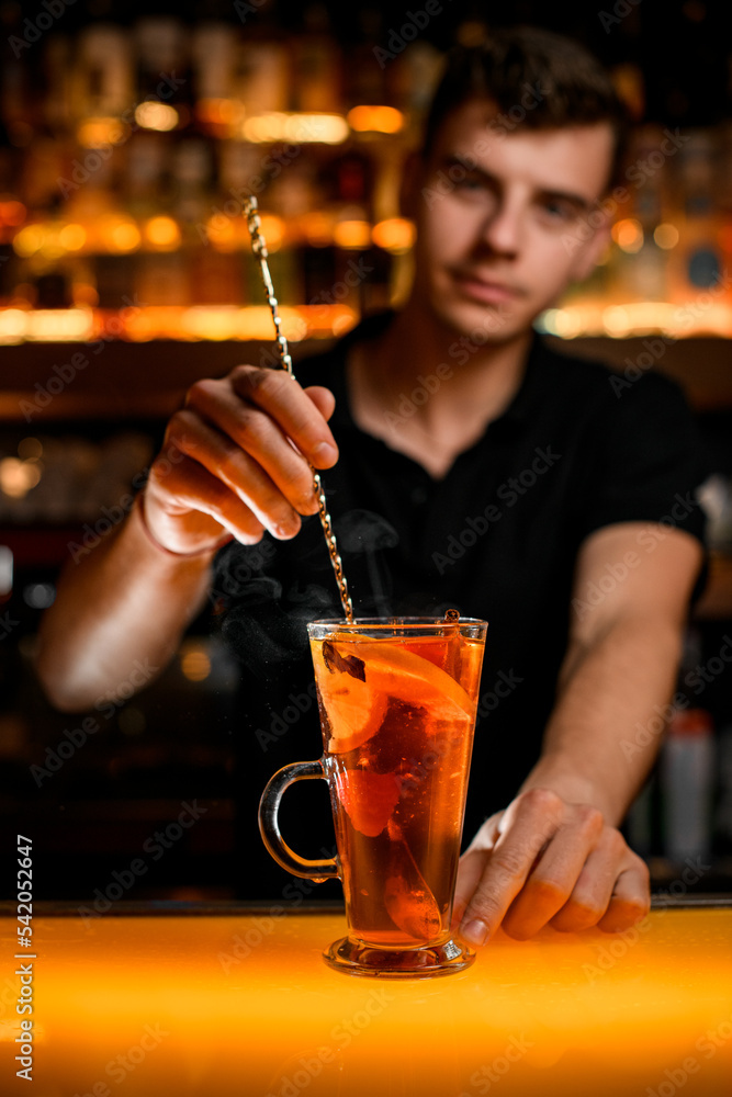 hand of bartender holds spoon and stirs drink with lemon in transparent cup on the bar
