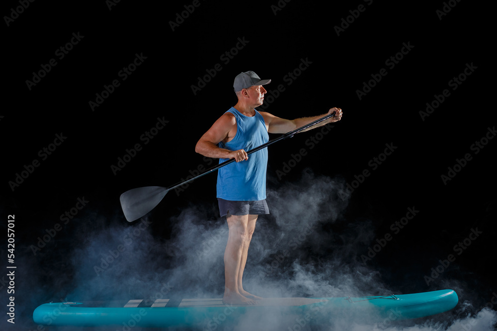 A man on a sub board with a paddle on a black background in the fog.
