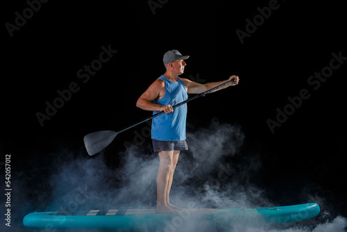 A man on a sub board with a paddle on a black background in the fog.