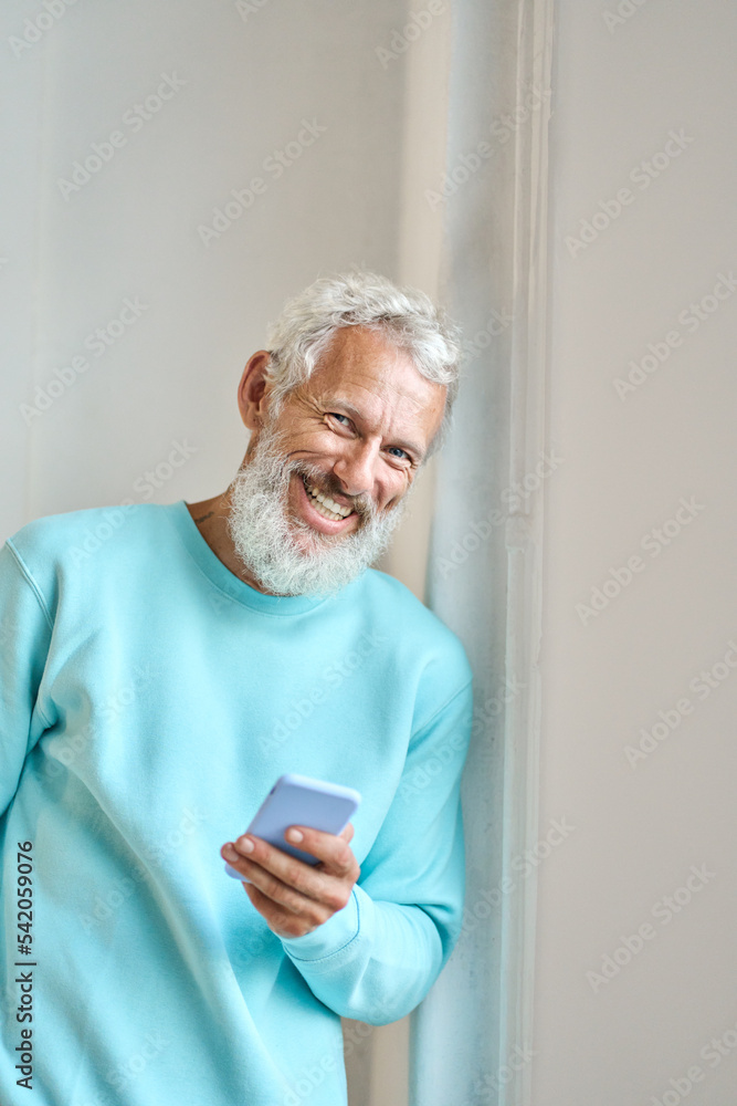 Happy smiling old senior middle aged bearded hipster man holding cell phone using cellphone modern smartphone device for chatting, making payments, e-learning standing at white wall. Happy smiling old senior middle aged bearded hipster man holding cell phone using cellphone modern smartphone device for chatting, making payments, e-learning standing at white wall.