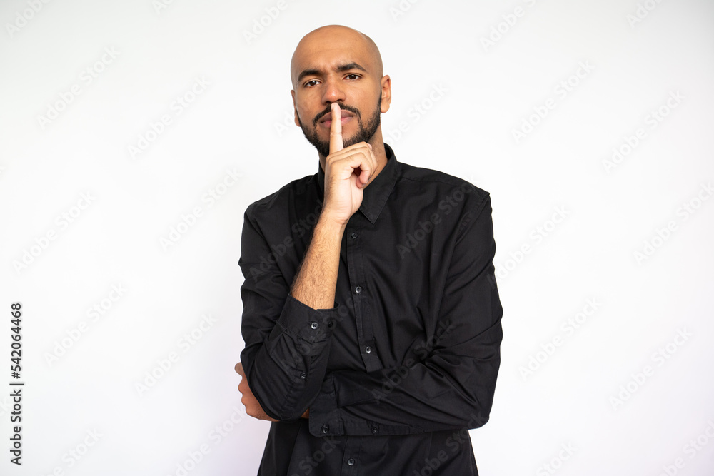 Portrait of confident young man standing with finger on lip against white background. Young bearded businessman wearing black shirt making silence or secret gesture. Secret concept Portrait of confident young man standing with finger on lip against white background. Young bearded businessman wearing black shirt making silence or secret gesture. Secret concept