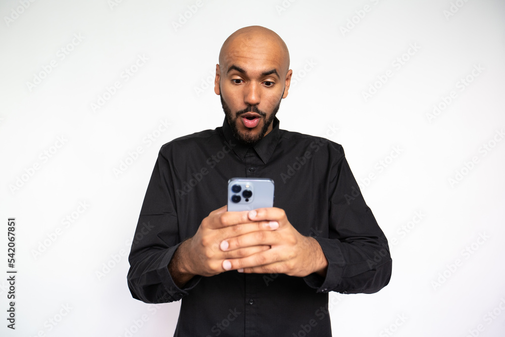 Portrait of astounded young man reading message on mobile phone against white background.Bearded businessman wearing black shirt surfing Internet on smartphone. Good news and mobile technology concept Portrait of astounded young man reading message on mobile phone against white background.Bearded businessman wearing black shirt surfing Internet on smartphone. Good news and mobile technology concept