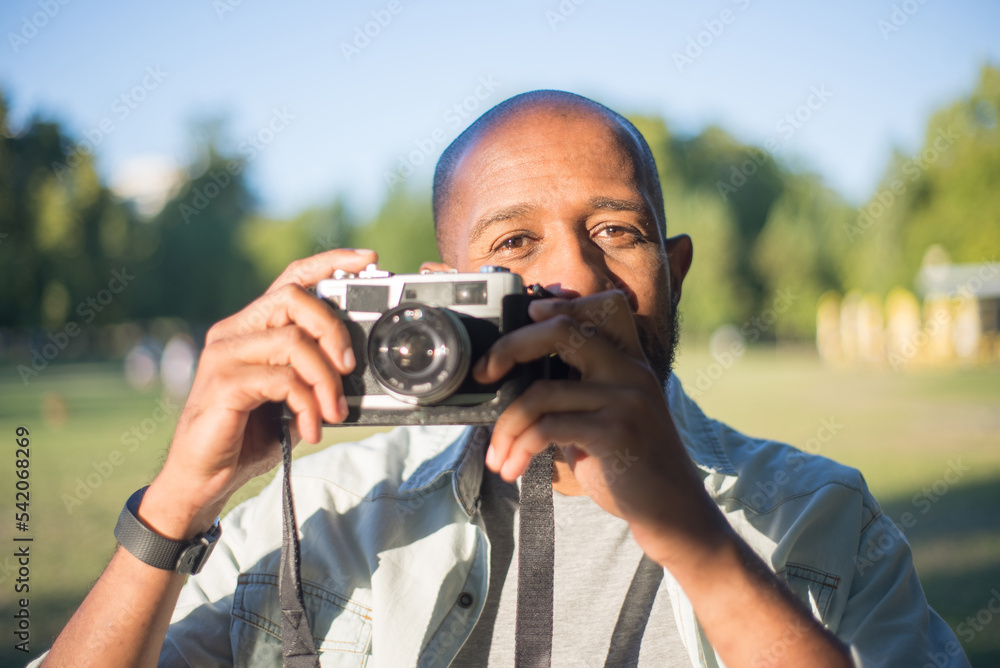 Close-up of happy African American man holding photo camera. Handsome bearded man standing in park looking at camera getting ready to take picture by himself. Leisure,hobby,modern technologies concept Close-up of happy African American man holding photo camera. Handsome bearded man standing in park looking at camera getting ready to take picture by himself. Leisure,hobby,modern technologies concept
