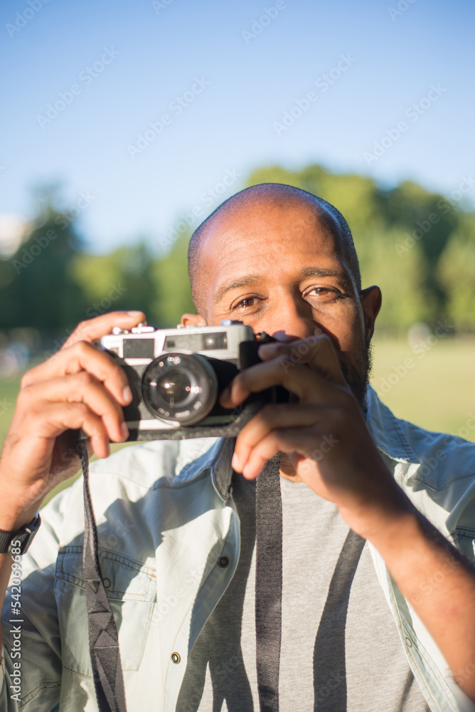 Close-up of serious African American man with photo camera. Concentrated brown-eyed man holding camera looking straight and getting ready to take photo. Hobby and modern technologies concept