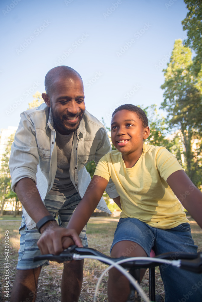 Careful African American man and son enjoying to ride bike. Happy father walking holding boy hand on bike handlebar helping to keep balance both happy having fun. Parents love, summer activity concept