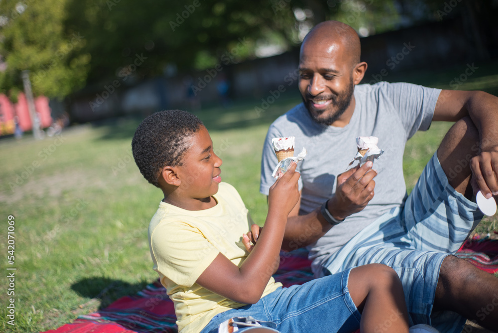 Happy daddy and little son spending time together. Smiling African American man and cute boy sitting on ground holding ice-cream cones talking in summer park. Parenting and leisure concept