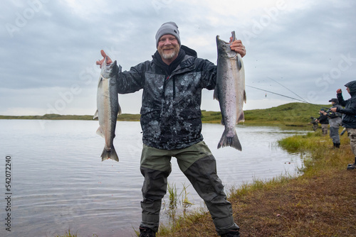Happy man proudly holds two large salmon fish caught on Egegik river in Alaska as other fishermen catch fish in the background photo
