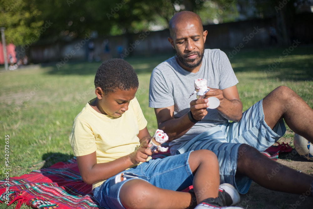 Portrait of happy man and boy resting together on ground. African American father and his kid sitting on blanket in summer park concentrated on unwrapping ice cream cones. Resting and leisure concept