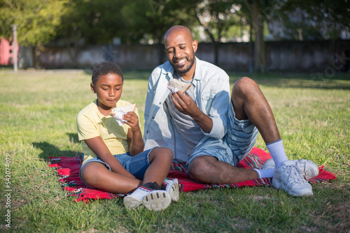 Portrait of young African American dad and kid holding sandwiches. Joyful bearded man and boy with curly hair sitting on blanket looking at their sandwiches with appetite. Leisure and summer concept © KAMPUS