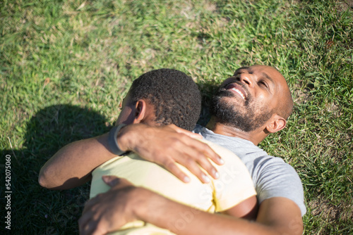 Close-up of loving African American father hugging his little son. Smiling man lying on grassy ground and boy with curly hair on his chest in summer. Parents love, care, time together concept © KAMPUS
