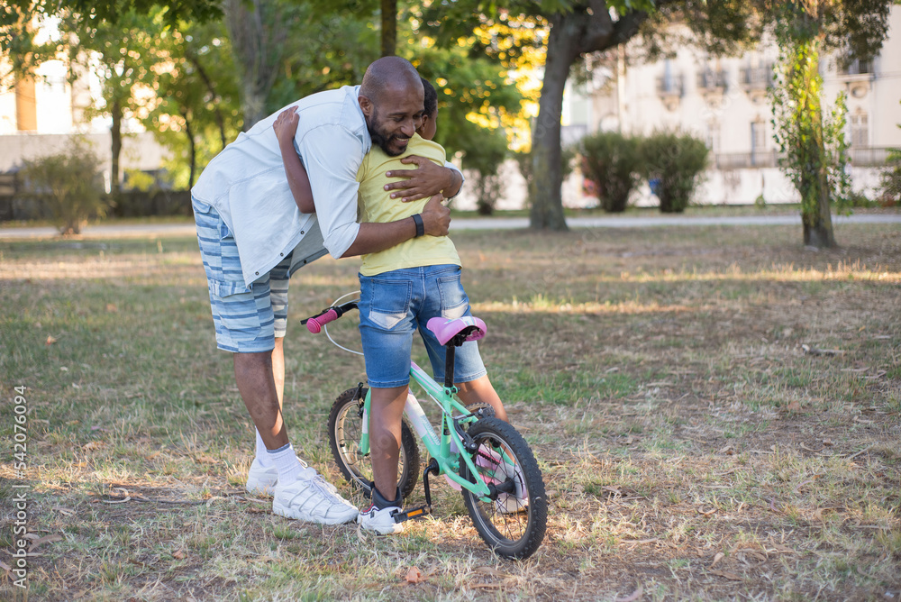 Obraz premium Portrait of happy African American dad and son training in park. Smiling man and boy with bike standing in city park and father hugging kid with love. Active summer leisure and parenthood concept