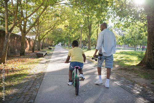 Wallpaper Mural Back view of African man and boy on shady alley in public park. Bearded dad walking holding skateboard looking at son and happy kid riding small bike. Spending time together and childhood concept Torontodigital.ca