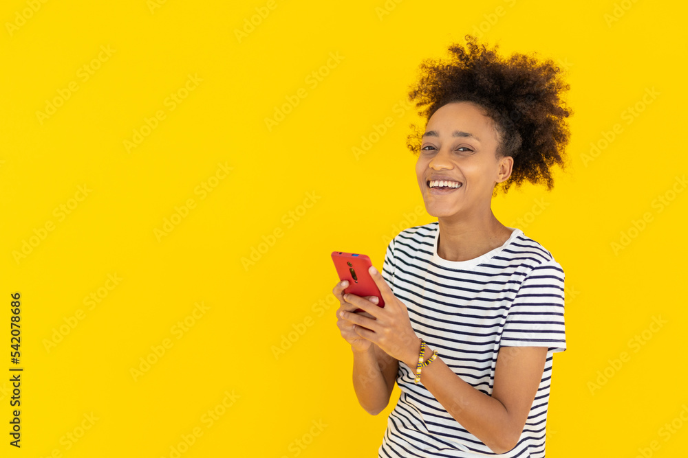 Happy young african woman smiling at camera using mobile phone against yellow background Happy young african woman smiling at camera using mobile phone against yellow background