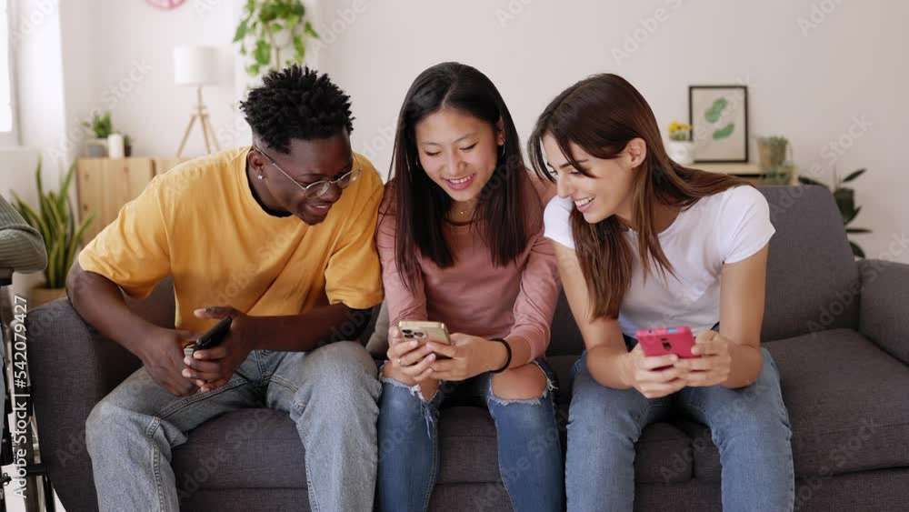 Three millennial teenager friends having fun using mobile phone sitting on sofa at home. Happy young people watching funny media content on social media app network. Three millennial teenager friends having fun using mobile phone sitting on sofa at home. Happy young people watching funny media content on social media app network.