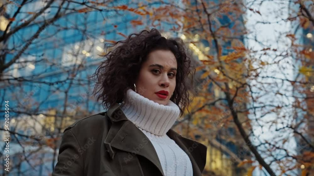 Slow motion portrait confident young woman with curly hair posing in autumn park near skyscrapers in downtown street looking at camera seductively, attractive female, urban style, evening city Slow motion portrait confident young woman with curly hair posing in autumn park near skyscrapers in downtown street looking at camera seductively, attractive female, urban style, evening city