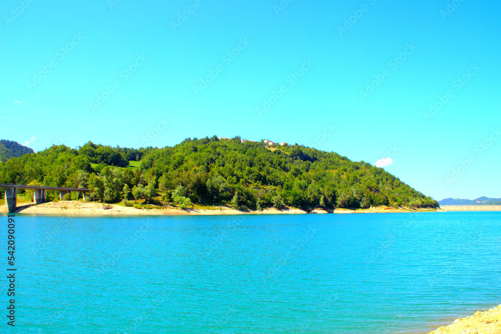 View on the shores of Gerosa Lake with its wooly silky blue waters delimited by a dam in the distance, a hill full of green vegetation and a bridge connecting through the wilderness on a summer day