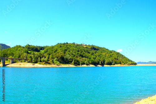 View on the shores of Gerosa Lake with its wooly silky blue waters delimited by a dam in the distance, a hill full of green vegetation and a bridge connecting through the wilderness on a summer day