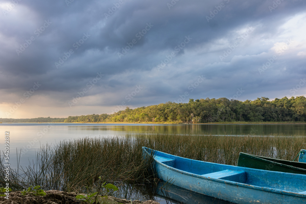 Blue canoe on shore with marshes and fresh water lagoon surrounded by tropical green jungle with light reflection and cloudy sky at sunset