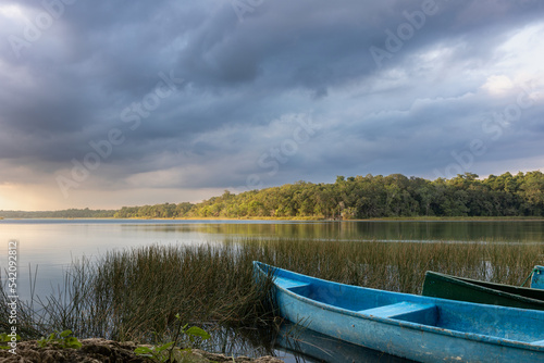 Blue canoe on shore with marshes and fresh water lagoon surrounded by tropical green jungle with light reflection and cloudy sky at sunset © Cualera