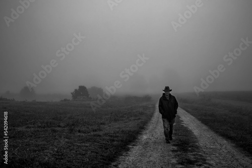 A moody vibe as a man saunters along a dirt road towards the viewer on a calm foggy morning.