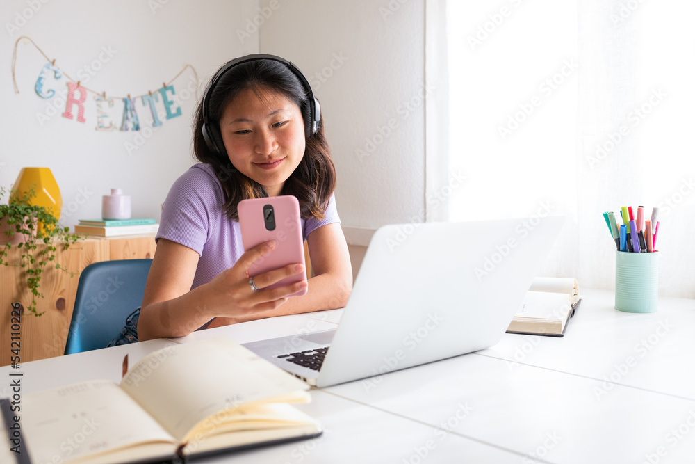 Asian female college student at home using laptop , phone and headphones while studying. Reading text message. Asian female college student at home using laptop , phone and headphones while studying. Reading text message.