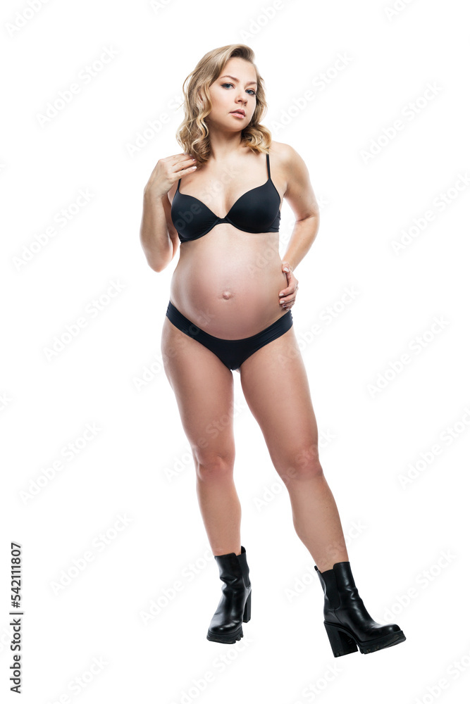 Fototapeta premium Young blonde pregnant woman in black lingerie and boots. Taking care of yourself while expecting a baby. Full height. Isolated on white background. Vertical.