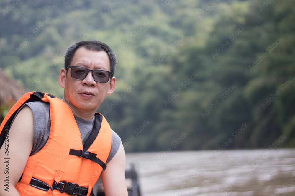 A man wearing a life jacket while waiting to swim, looking at the camera sitting on a raft by the water. A man wearing a life jacket while waiting to swim, looking at the camera sitting on a raft by the water.