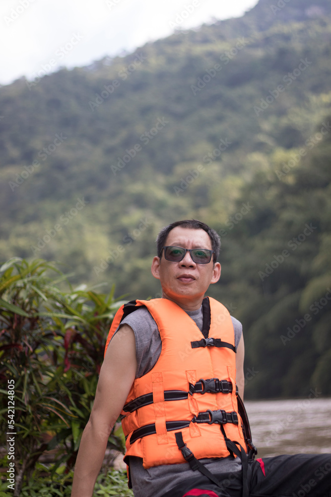 A man wearing a life jacket while waiting to swim, looking at the camera sitting on a raft by the water. A man wearing a life jacket while waiting to swim, looking at the camera sitting on a raft by the water.