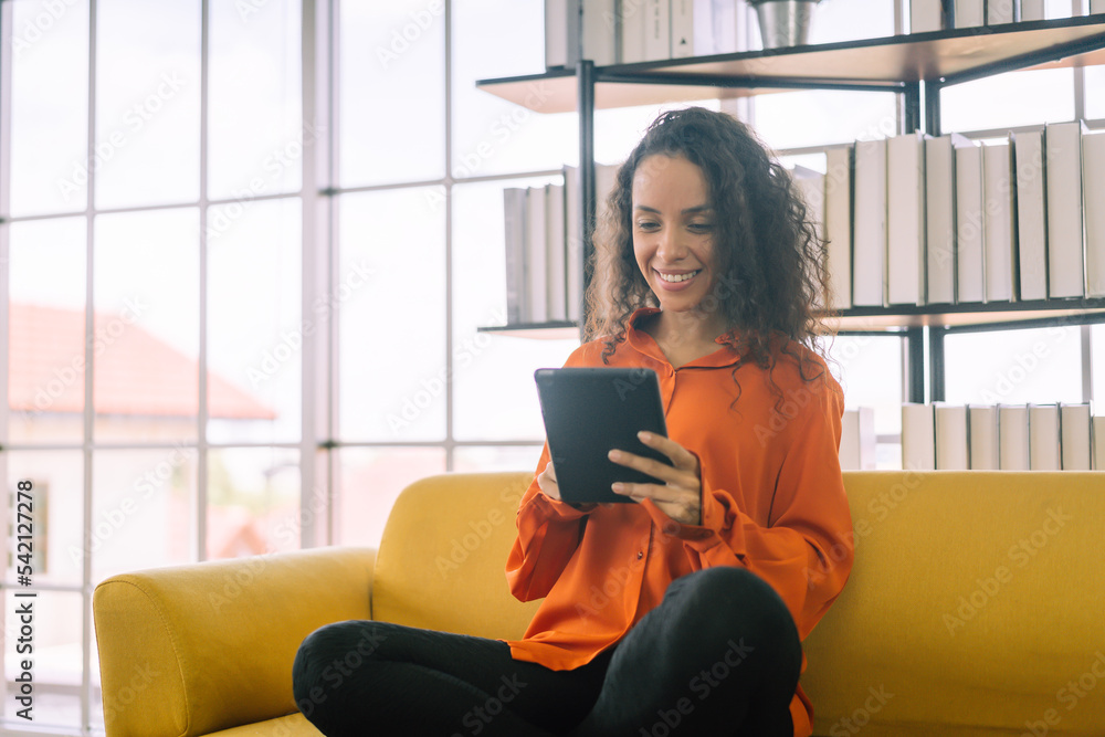 Latin woman is using a digital tablet and smiling while sitting on sofa Latin woman is using a digital tablet and smiling while sitting on sofa