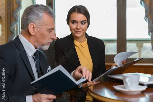 Young businesswoman showing documents to adult businessman while drinking coffee in luxury hotel