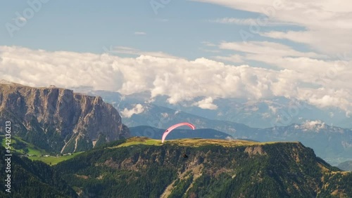 Group of paragliders flies over green forested highland in rocky mountains. High cloudy peaks of Alps under blue sky on summer day aerial view
