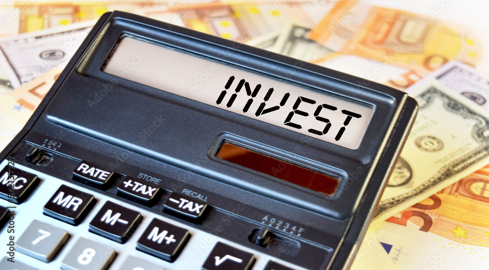 INVEST text written on a calculator next to the background of euro banknotes. Finance or business concept. INVEST text written on a calculator next to the background of euro banknotes. Finance or business concept.