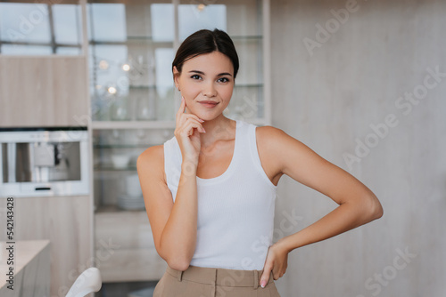 Playful young asian woman in white t-shirt and beige pants over blurry living room. Pretty Korean model touching her face with perfect skin looks at camera smiling. Fit female home. Beauty and health.