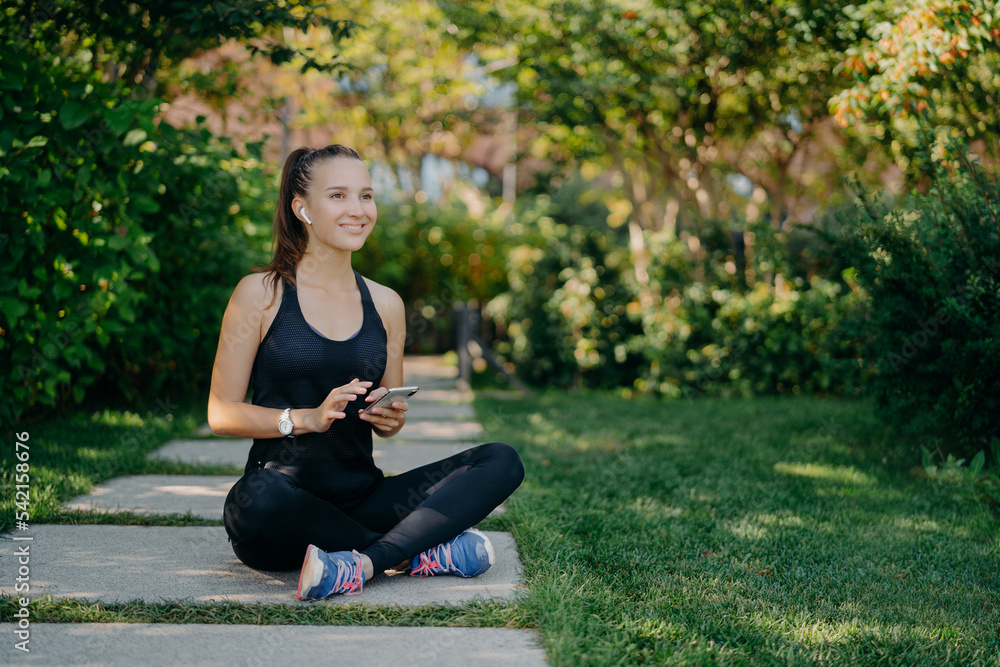 Cheerful female runner poses in lotus pose outdoor dressed in sportswear uses mobile phone during rest after jog downloads music songs in player prepares for morning workout chooses audio records Cheerful female runner poses in lotus pose outdoor dressed in sportswear uses mobile phone during rest after jog downloads music songs in player prepares for morning workout chooses audio records