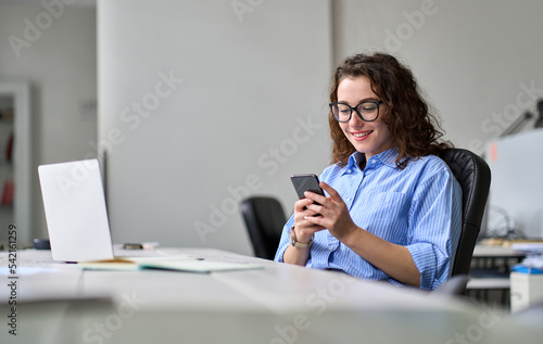 Young happy business woman entrepreneur looking at smartphone using cellphone mobile cell corporate tech, professional female manager working in office typing on mobile cell phone sitting at desk.