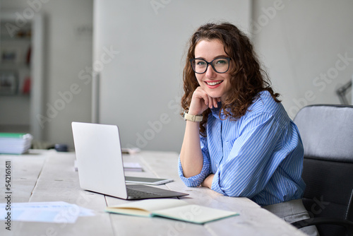 Young happy business woman company employee sitting at desk working on laptop. Smiling female professional entrepreneur worker using computer in corporate modern office looking at camera. Portrait. photo