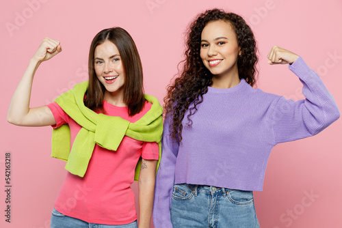 Young two friends women wears green purple shirts looking camera together show biceps muscles on hand demonstrating strength power isolated on pastel plain light pink color background studio portrait.