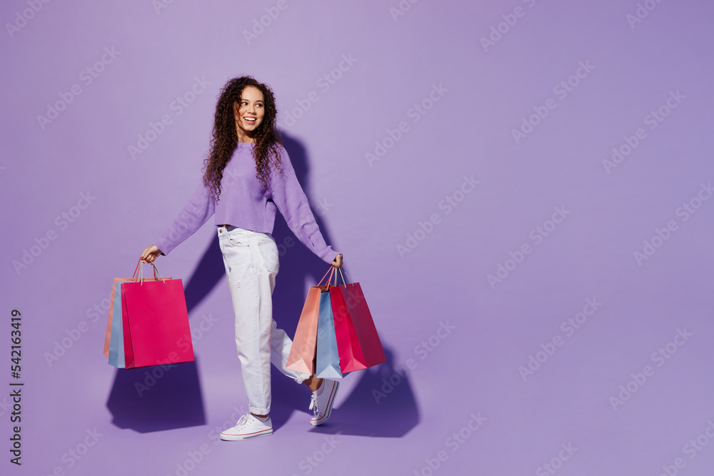 Full body side view young woman wear pullover hold in hand paper package bags after shopping walking go look aside isolated on plain pastel light purple background. Black Friday sale buy day concept. Full body side view young woman wear pullover hold in hand paper package bags after shopping walking go look aside isolated on plain pastel light purple background. Black Friday sale buy day concept.