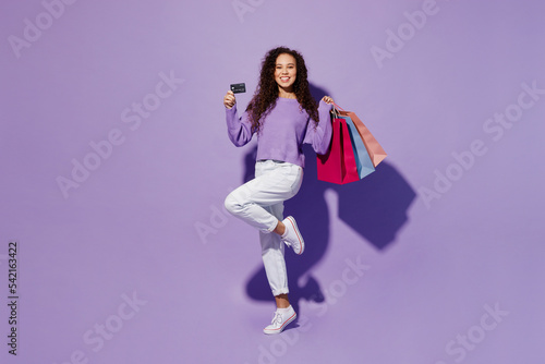 Full body side view happy young woman wear pullover hold in hand paper package bags after shopping credit bank card isolated on plain pastel light purple background. Black Friday sale buy day concept.