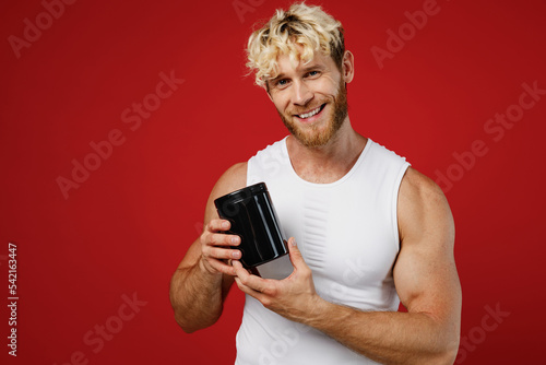 Young happy strong sporty sportsman toned man wear white clothes spend time in home gym hold black bottle of sport protein supplement isolated on plain red background. Workout sport fit body concept.