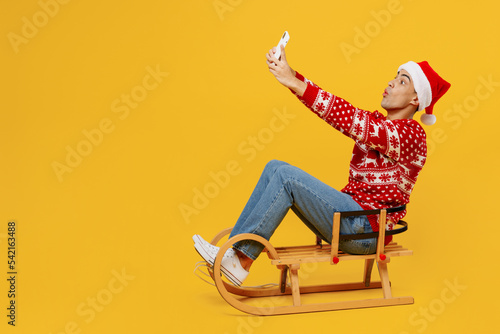 Full size merry young man wear red knitted Christmas sweater Santa hat posing sledding doing selfie shot on mobile cell phone isolated on plain yellow background. Happy New Year 2023 holiday concept.