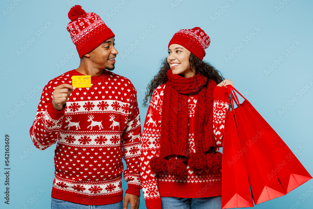 Merry young couple two man woman wear red Christmas sweater Santa hat posing hat hold shopping package bag credit bank card isolated on plain pastel blue background Black Friday sale buy day concept.