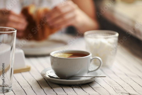 Woman eating croissant at table, focus on cup of aromatic tea with lemon