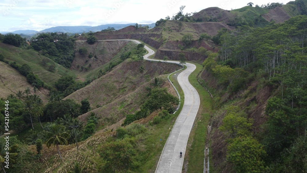 Aerial view of curvy road surrounded by high hills and mountains with green lush tree forest